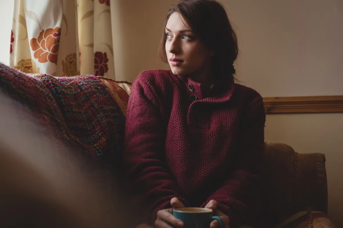 Eine junge Frau sitzt nachdenklich mit Kaffeetasse auf dem Sofa.